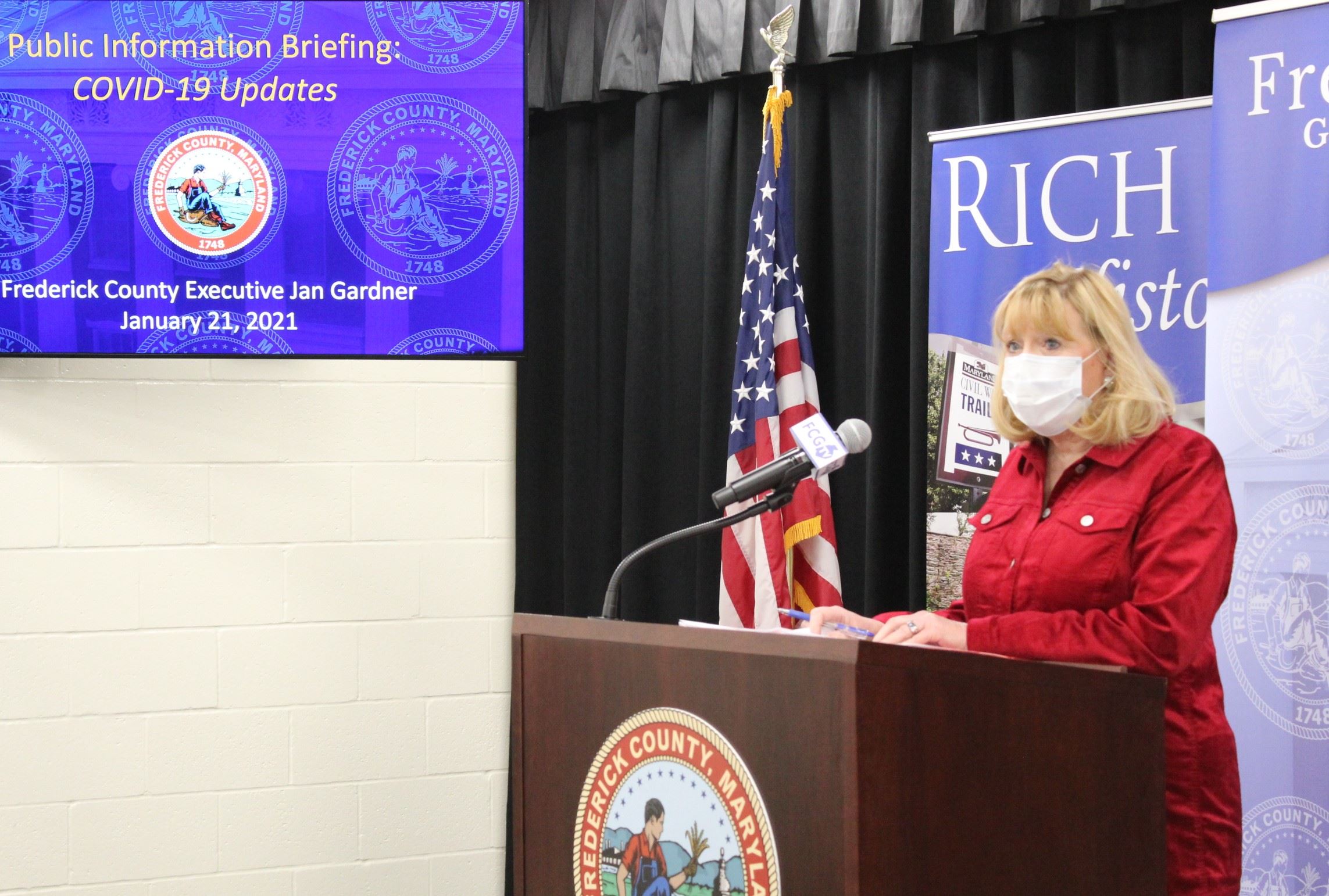 County Executive Gardner Stands at a Lectern. Beside her is a monitor with the words Covid-19 Update