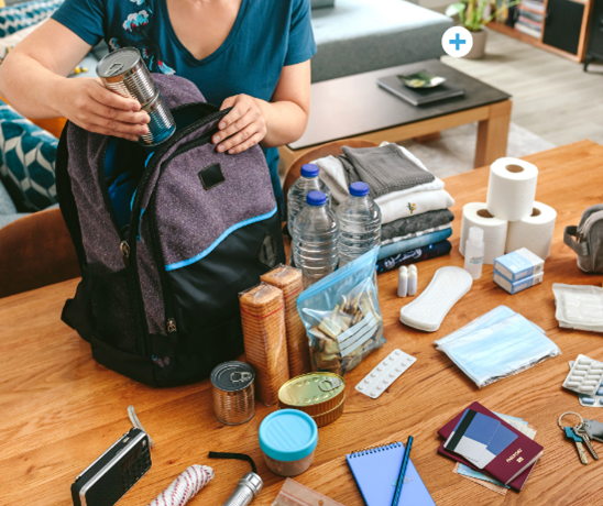 A woman holding a book bag open with one hand and using the other to put items in it from a table. 