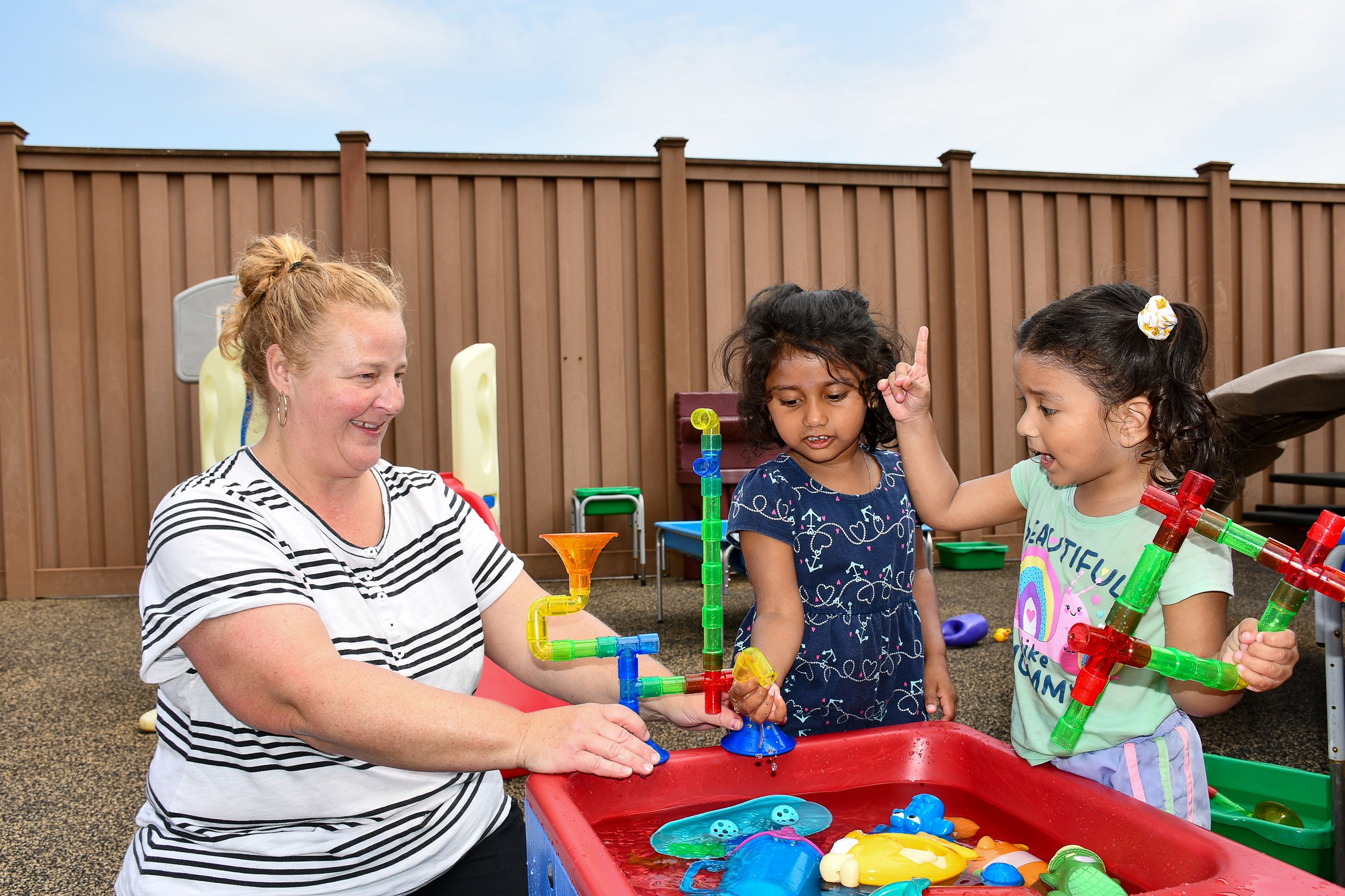 A teacher smiles while playing at a water table with two young children.