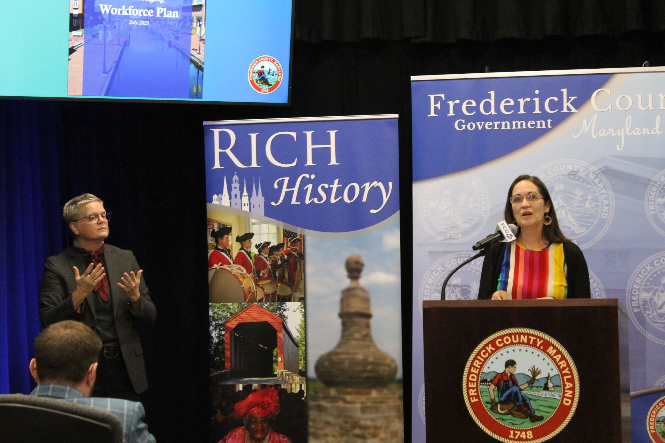 A woman standing inside at a microphone, with a sign language interpreter to her side
